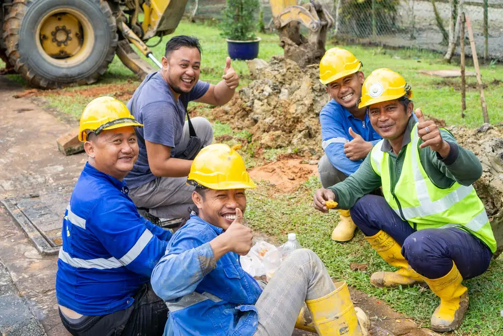 Equipe de obra com capacetes e coletes, sentada ao ar livre e fazendo sinal de joia para a câmera