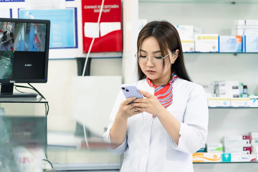Farmacêutica de jaleco usando o celular atrás do balcão de uma farmácia, com medicamentos nas prateleiras