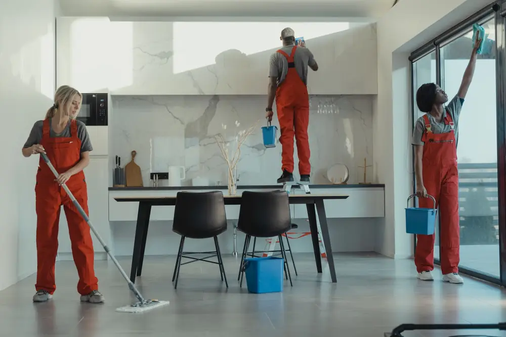Equipe de limpeza de macacão vermelho passando pano no chão e limpando vidros em uma cozinha ampla
