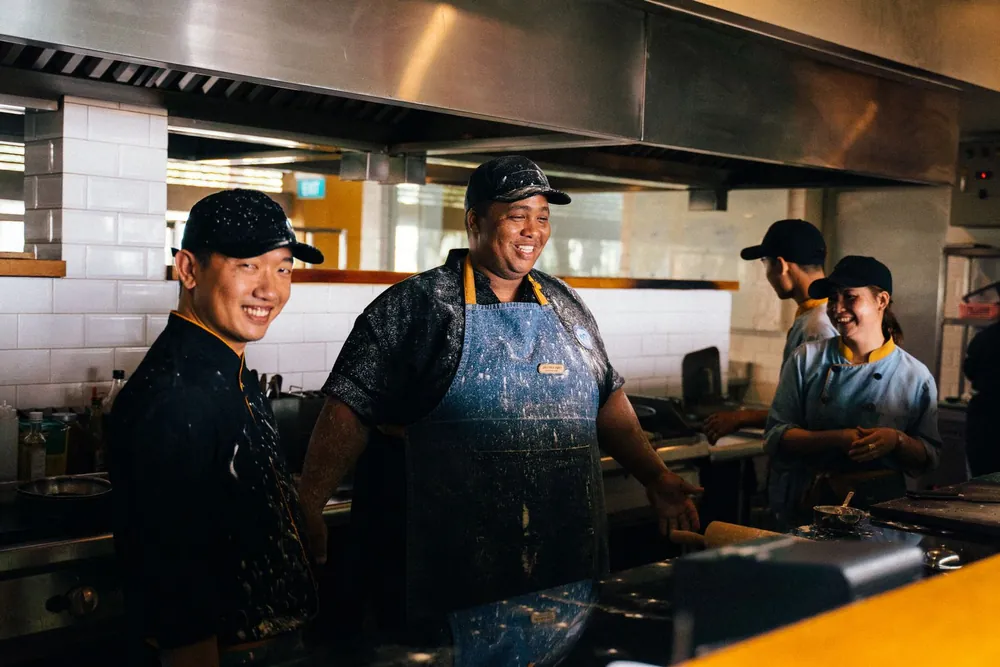 Cozinheiros sorrindo em uma cozinha de restaurante, com outros funcionários trabalhando ao fundo